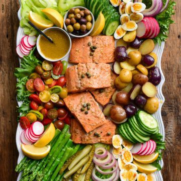 A large platter of a salmon Niçoise salad. Five pieces of salmon are centered in the platter and are surrounded by various vegetables, eggs and pickles.