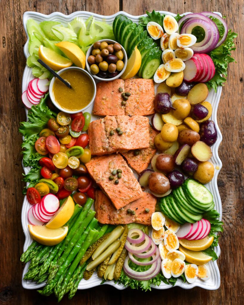 A large platter of a Niçoise Salad with Salmon. Five pieces of salmon are centered in the platter and are surrounded by various vegetables, eggs and pickles.