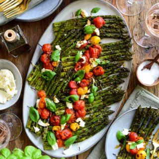 An oval platter with grilled asparagus 'rafts' topped with roasted tomatoes, mozzarella, basil and balsamic vinegar. This platter is surrounded by a platted portion and more ingredients.
