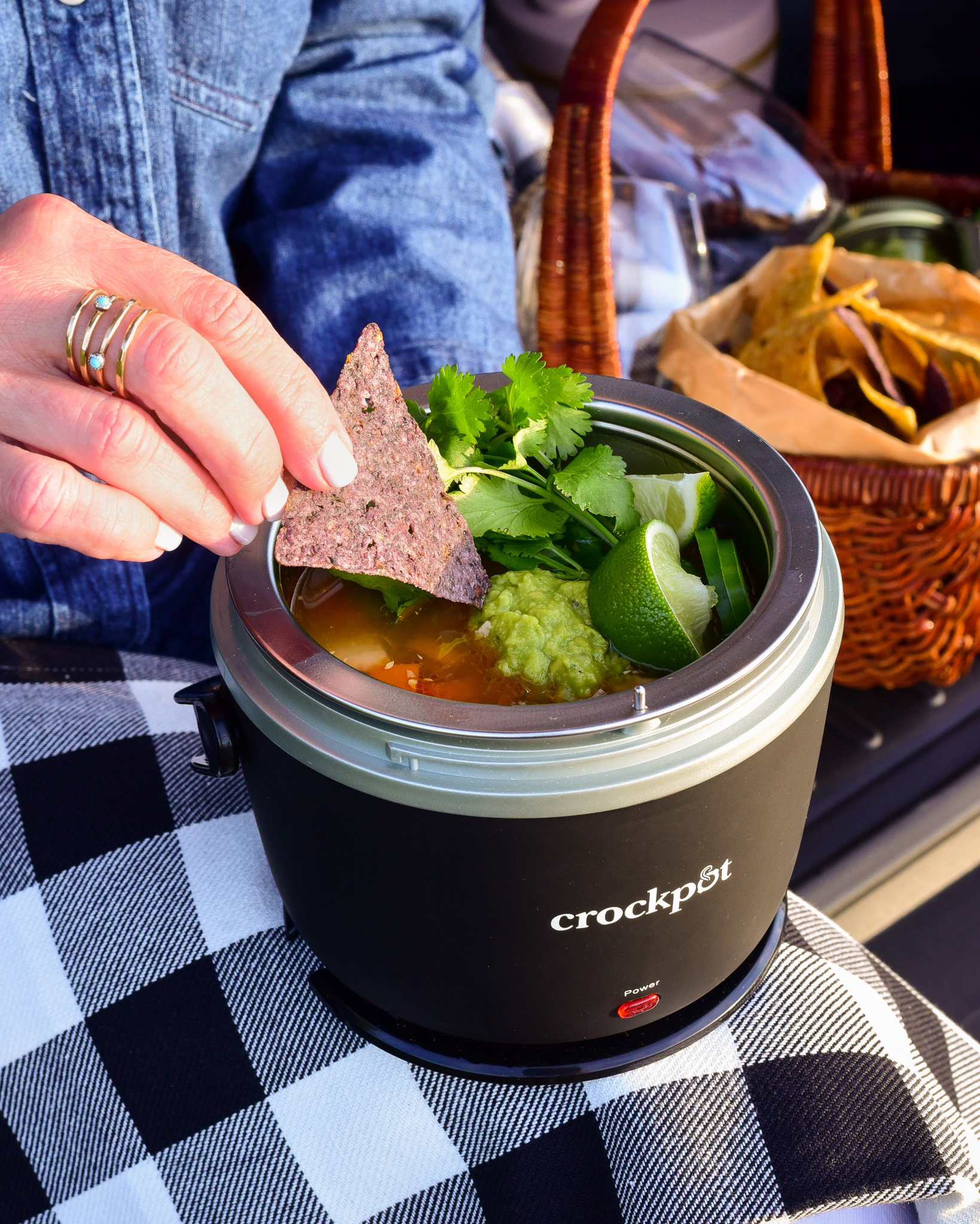 A woman's hand is dipping a nacho in her mini black portable crockpot on her lap. 