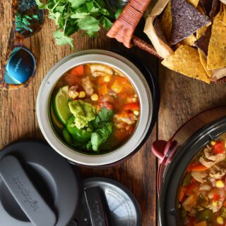 A twenty ounce mini lunch crock if filled with a bright and colourful chicken chili and is ready to be transported on a road trip along with a picnic basket.