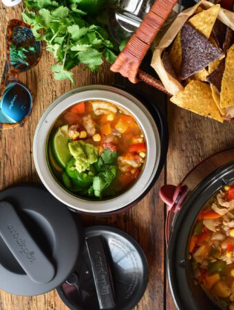 A twenty ounce mini lunch crock if filled with a bright and colourful chicken chili and is ready to be transported on a road trip along with a picnic basket.