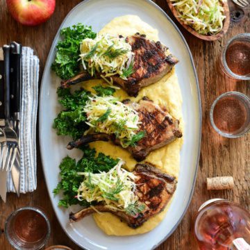 An oval platter with three pork chops topped with apple slaw on a creamy bed of polenta.
