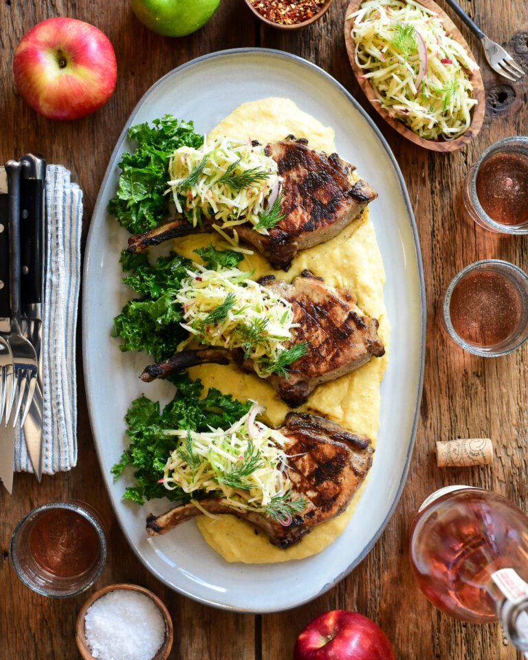 An oval platter with three pork chops topped with apple slaw on a creamy bed of polenta.