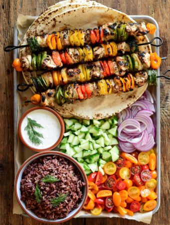 A pan with four chicken and vegetable skewers on top of a stack of large pitas. Served with rice, tzatziki, chopped cucumber, tomatoes and onions.