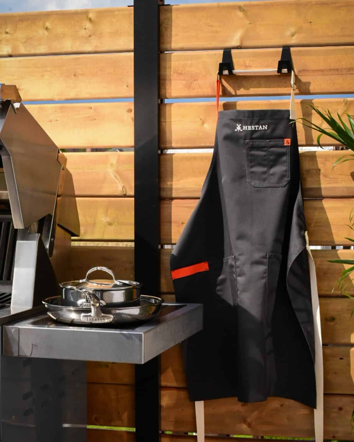 An apron is hanging outdoors beside a grill with pots and pans.
