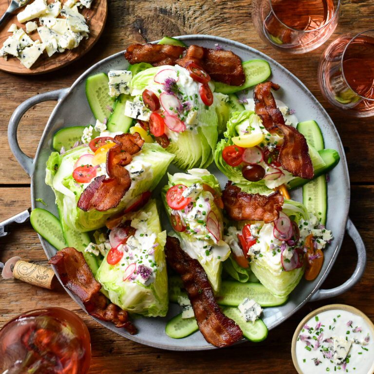 A round platter with six wedge salads topped with dressing, cheese, tomatoes and a strip of bacon. Served with rose wine.
