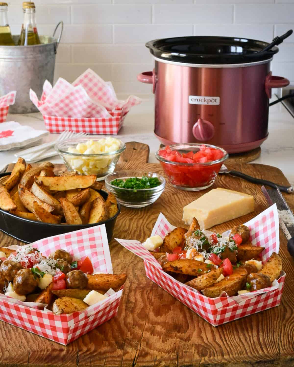 Two red and white check lined paper bowls are filled with poutine in the foreground. Potato wedges, poutine toppings and a copper slow cooker are in the background.