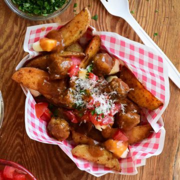 A top down view of poutine with meatballs. A red and white check snack bowl if filled with potato wedges and cheese curds which are covered in gravy and meatballs.