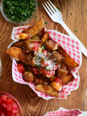 A top down view of poutine with meatballs. A red and white check snack bowl if filled with potato wedges and cheese curds which are covered in gravy and meatballs.