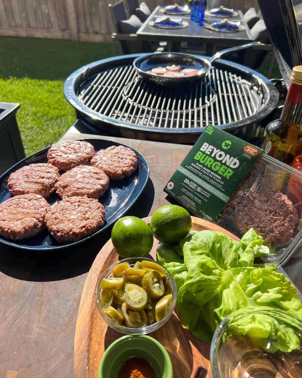 Burger patties and toppings are beside a bbq ready to be cooked.