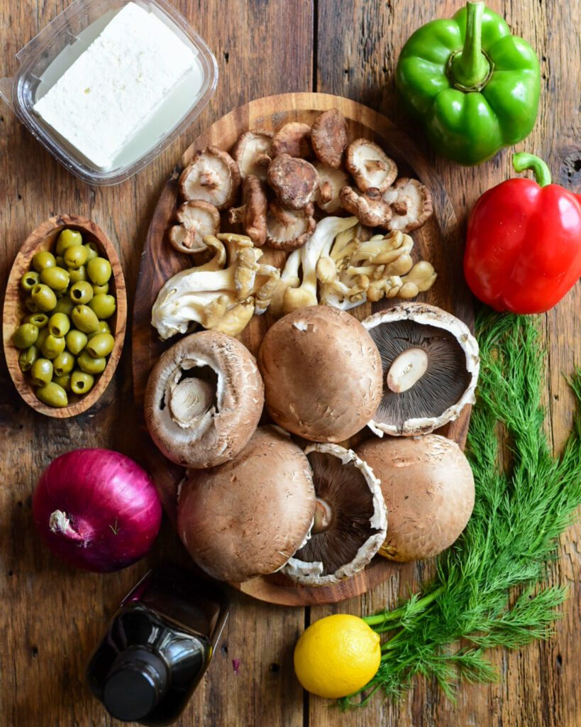A wooden plate with three different varieties of mushrooms. Peppers, onions, olives, dill, lemon and feta surround this plate.