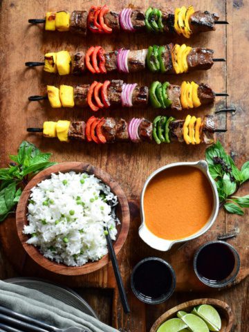 Four grilled beef skewers with alternating pieces of mango, peppers and onions on a board with a bowl of Thai curry sauce and a bowl of rice on a wooden board.