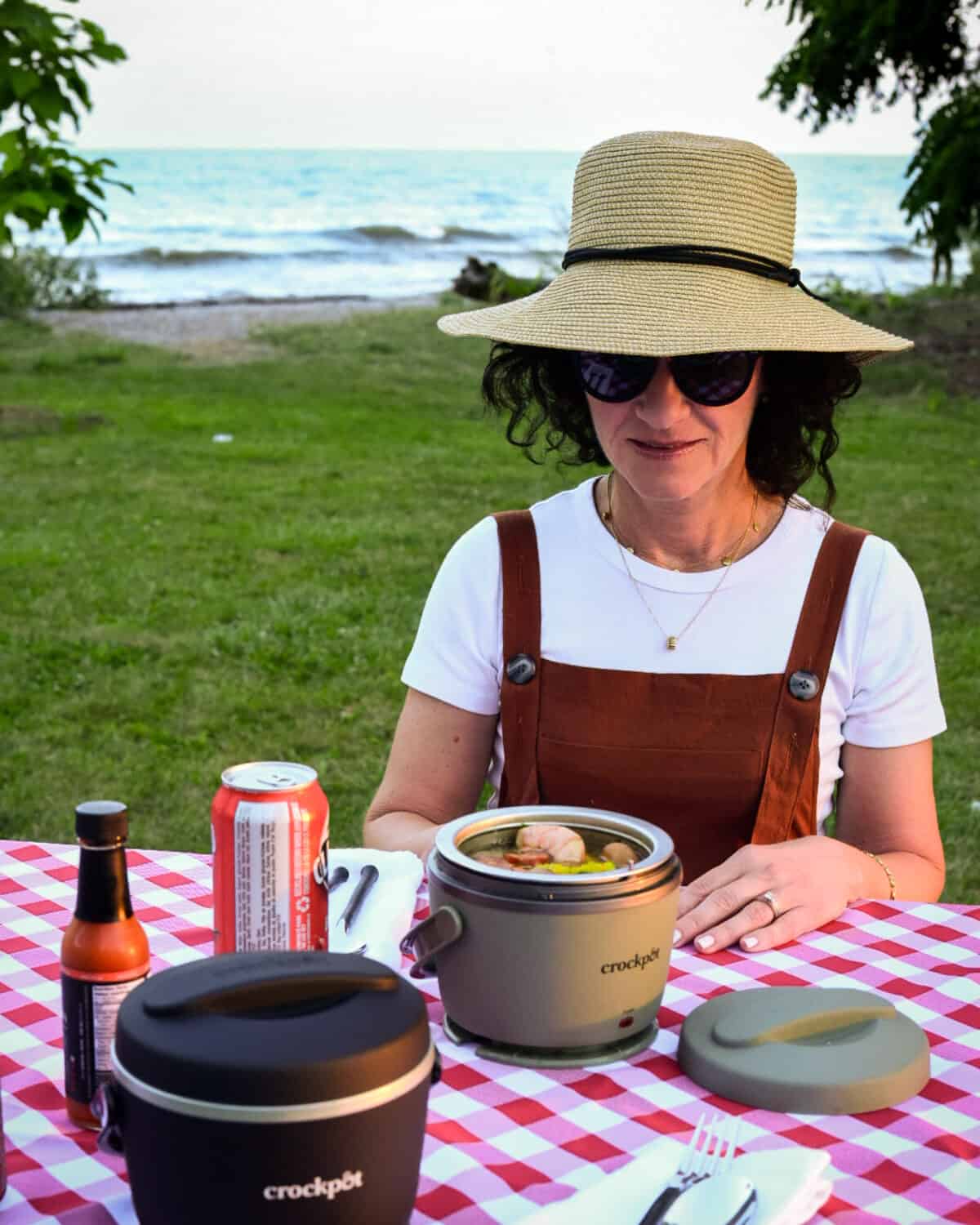 A woman wearing a straw hat sitting at picnic table with a red and white checkered tablecloth about to eat from her lunch crock pot.