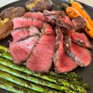 Several pieces of a medium rare steak fanned on a plate with grilled asparagus, potatoes and carrots.
