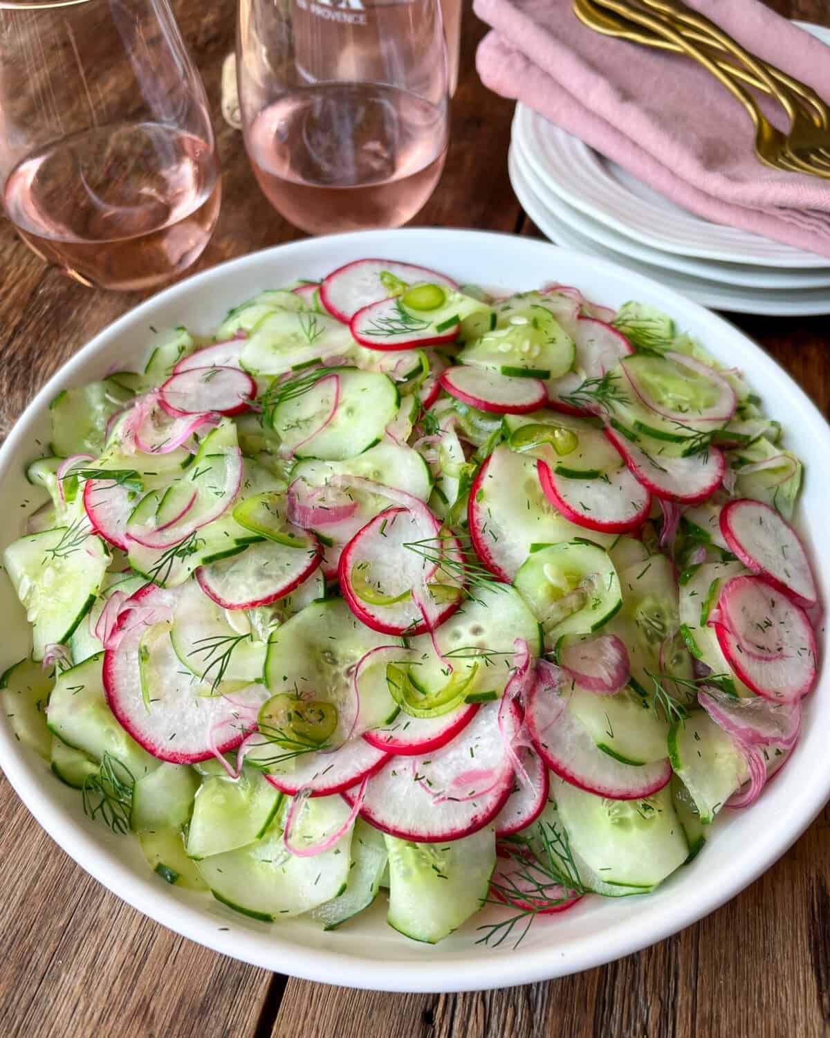 A side view of a salad of cucumbers, radishes, and jalapeños. A stack plates with pink napkins and gold cutlery and two glasses of rose wine are beside this bowl.