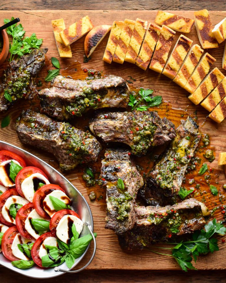 Seven grilled beef ribs with a green salsa on a board with grilled garlic toast and a Caprese salad.