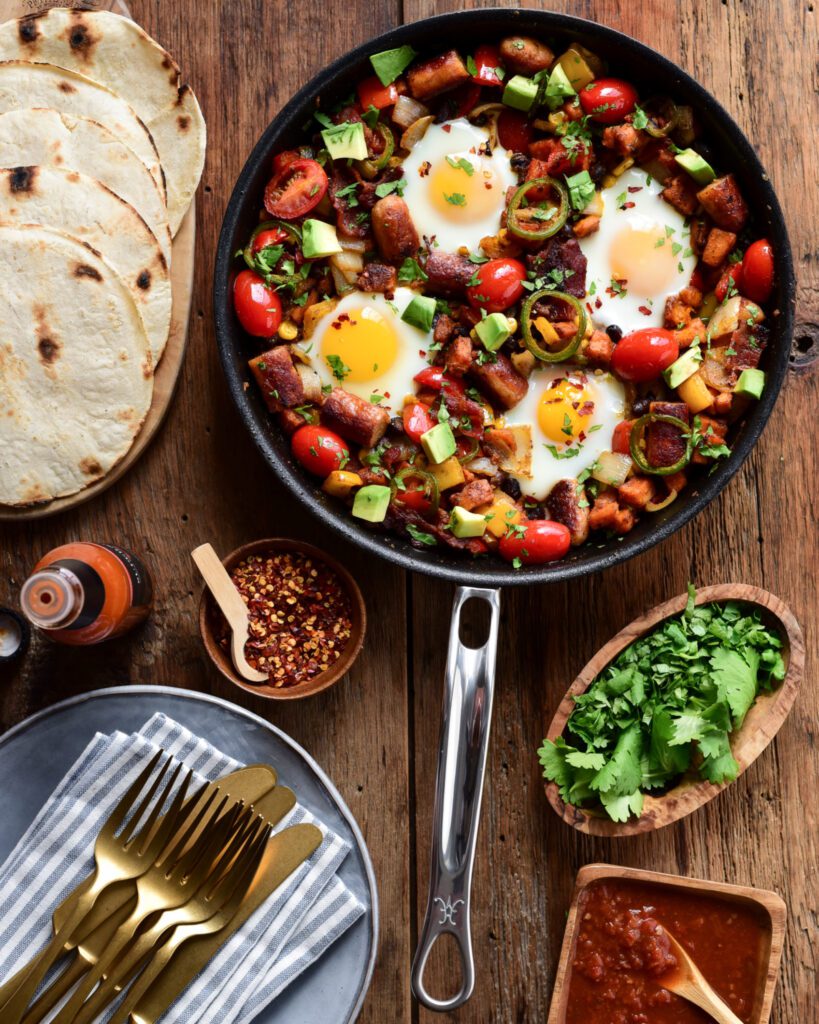 A one skillet hash with four eggs surrounded by plates, cutlery, hot sauce, cilantro and tortillas.