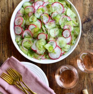 A salad of cucumbers, radishes, and jalapeños. A stack plates with pink napkins and gold cutlery and two glasses of rose wine are beside this bowl.