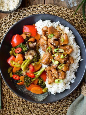 A black plate with rice and chicken on one side and bright cooked vegetables on the other side.