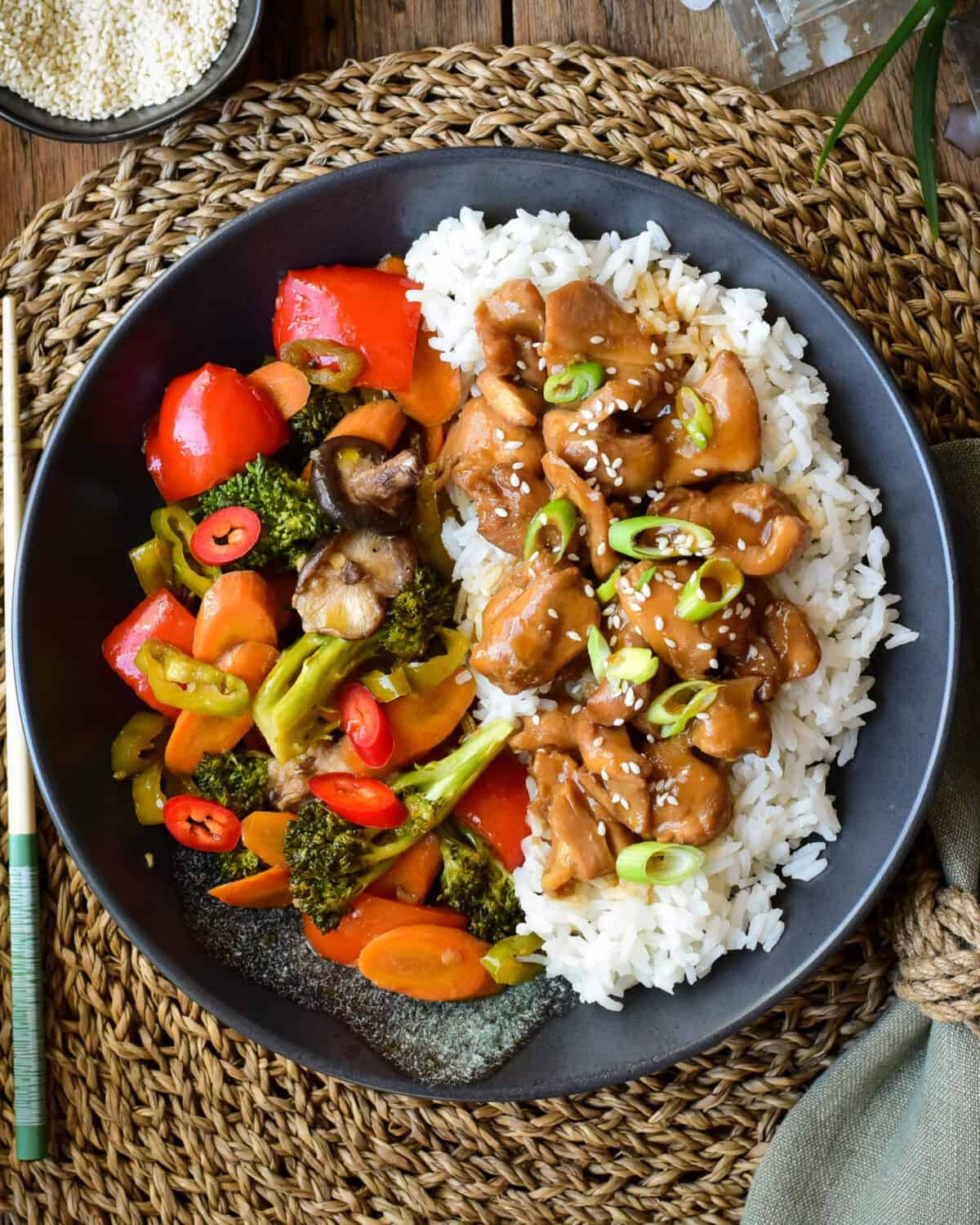 A black plate with rice and chicken on one side and bright cooked vegetables on the other side.