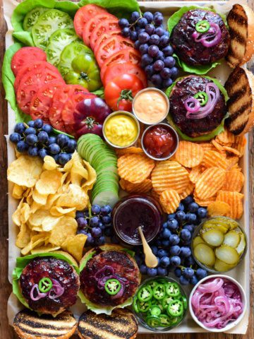 A top down image of a colourful tray with burgers, grape jam, blue grapes, tomatoes, chips and condiments.