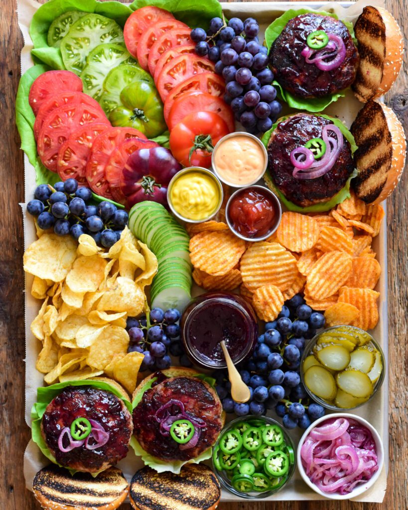 A top down image of a colourful tray with burgers, grape jam, blue grapes, tomatoes, chips and condiments.