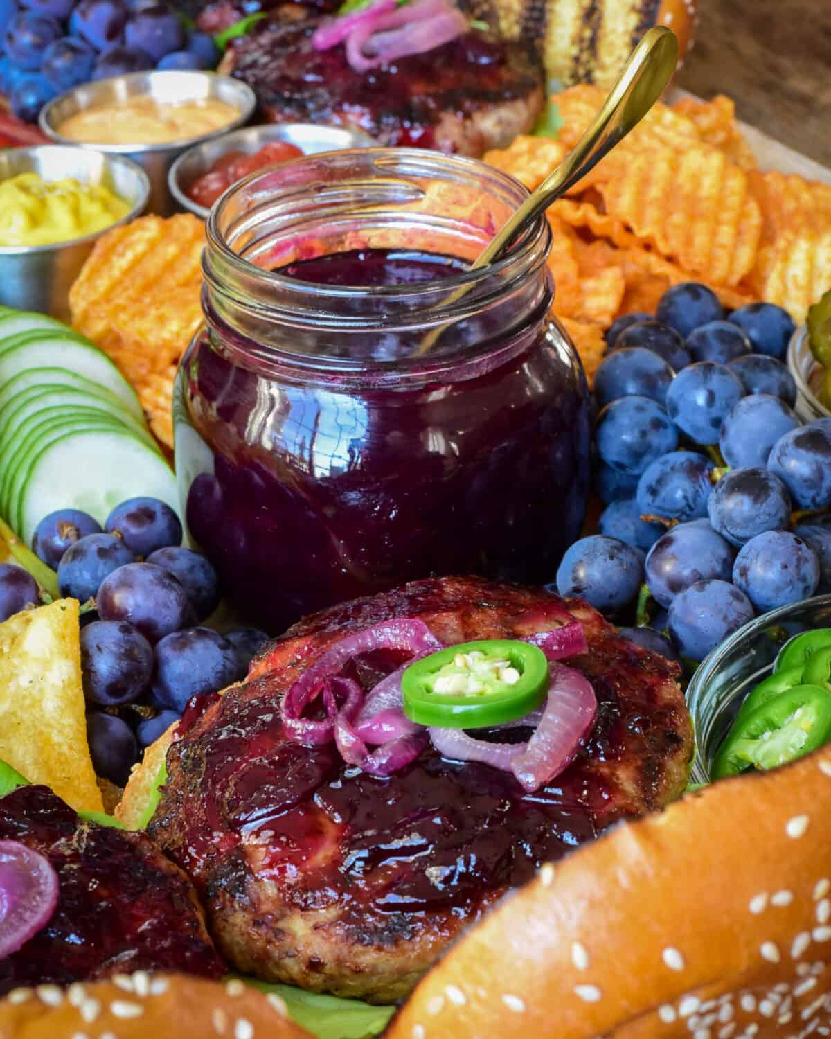 A jar of grape jam in a tray with a turkey burger, grapes, chips and cucumbers. 