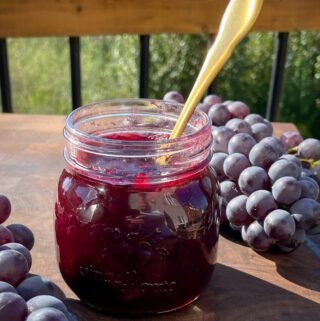 A jar of grape jam with with two bundles of grapes.