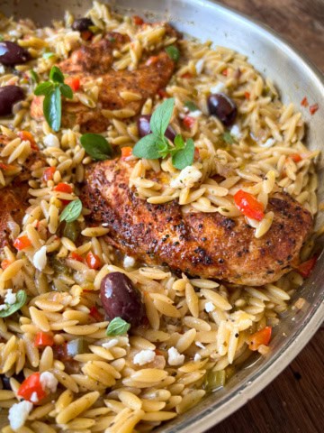 A skillet with chicken, orzo, olives and other ingredients.