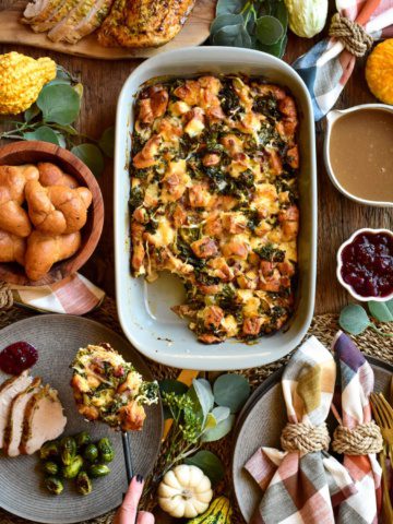 A Thanksgiving themed table setting with an oval dish of bread pudding in the center, with someone spooning out a portion onto a plate with turkey and Brussels sprouts.