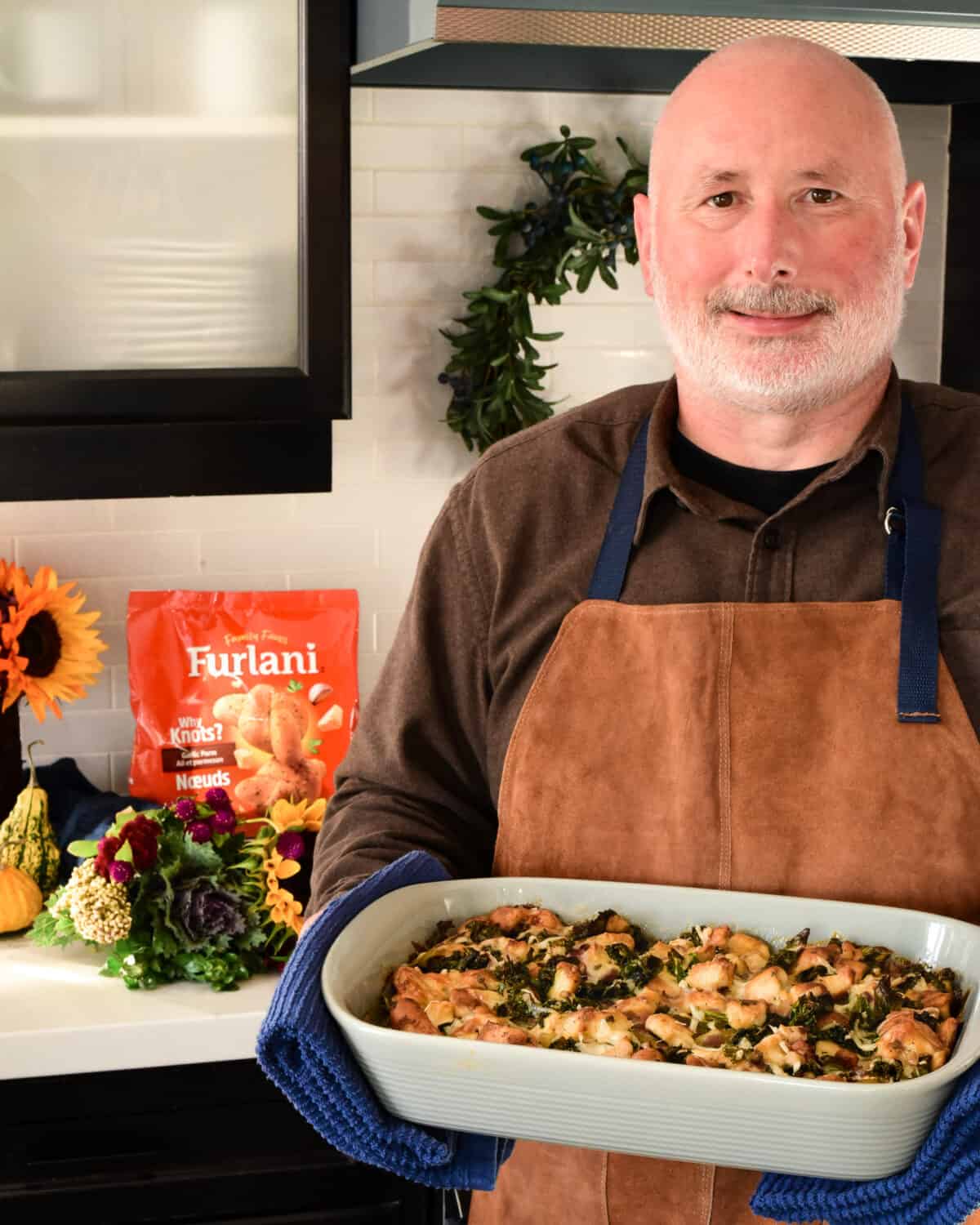 A man in suede apron holding the baking dish in his kitchen with fall flowers in the background.