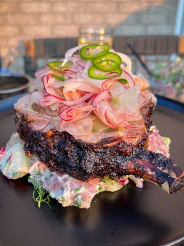 A rotisserie jerk pork chop is platted on a bed of potato salad and is topped with a apple fennel slaw and sliced jalapeños.