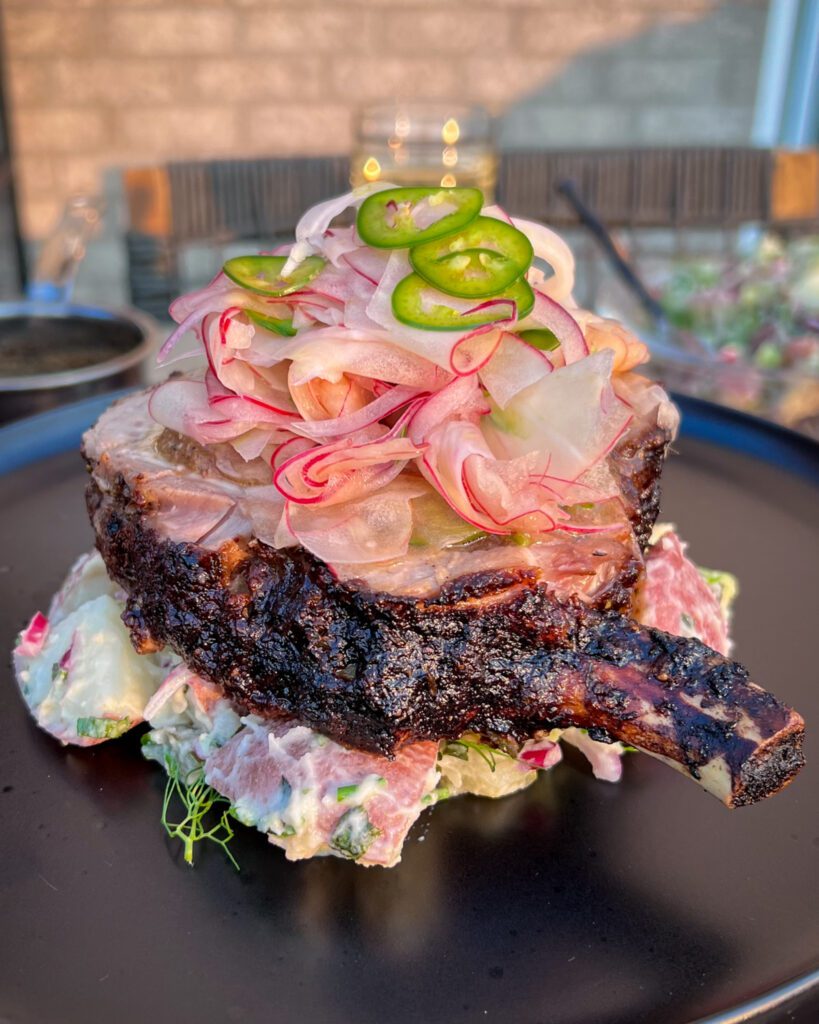 A rotisserie jerk pork chop is platted on a bed of potato salad and is topped with a apple fennel slaw and sliced jalapeños. 