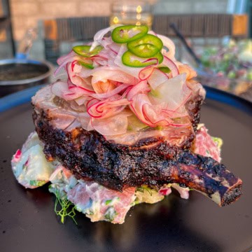 A thick bone-in pork rotisserie jerk pork chop sits on a bed of potato salad, topped with a slaw and other vegetables.