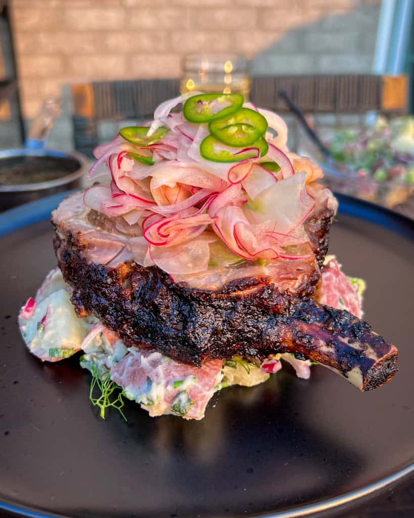 A thick bone-in pork rotisserie jerk pork chop sits on a bed of potato salad, topped with a slaw and other vegetables.