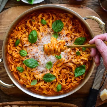 A sauté pan with homemade fresh pasta topped with basil and Parmesan. Someone is twirling a forkful of pasta.