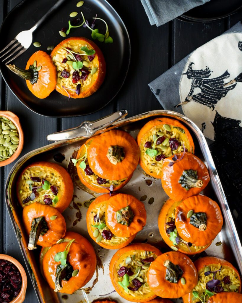 A top down image of baked mini stuffed pumpkins in a roaster with one on a plate. A Halloween witch tea towel is beside the roaster. 