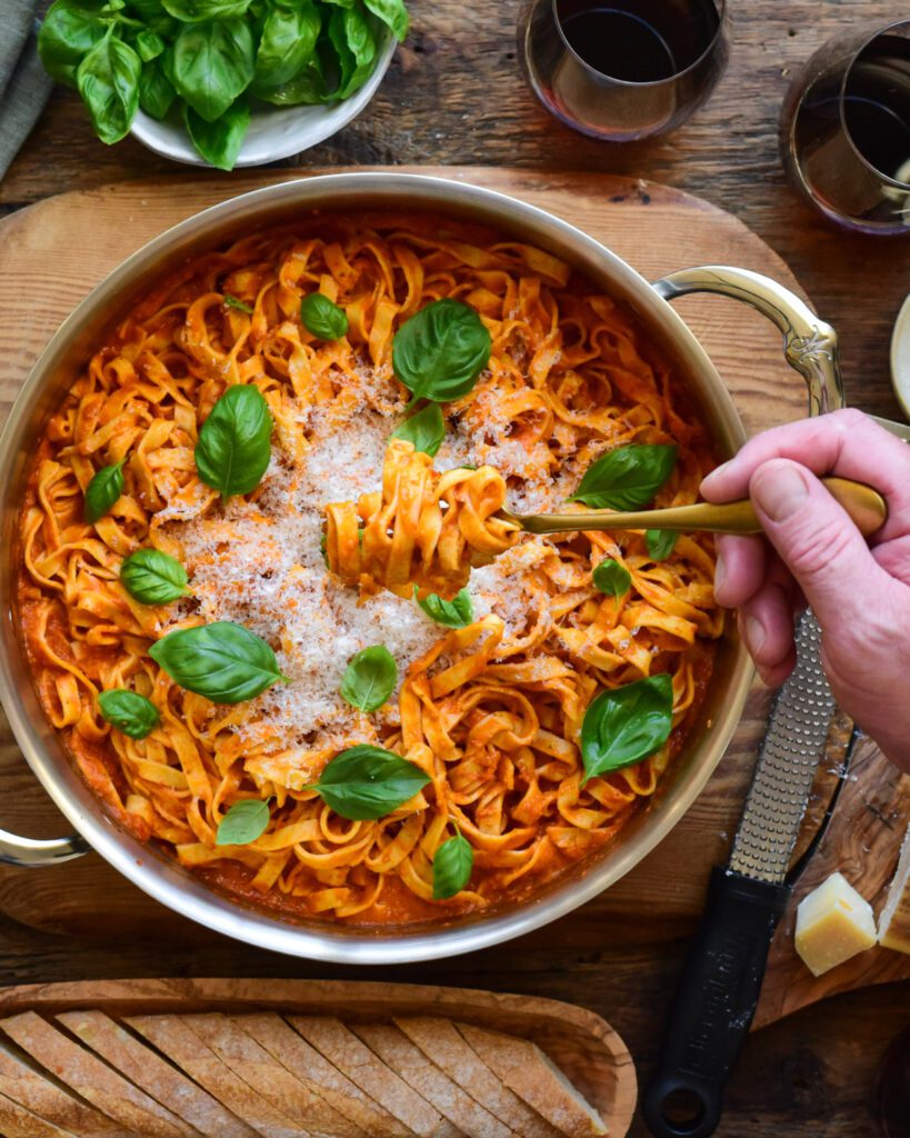 A pot of noodles in a tomato based pasta sauce with basil and shaved Parmesan. A twisted forkful of pasta is being held over this pot.  