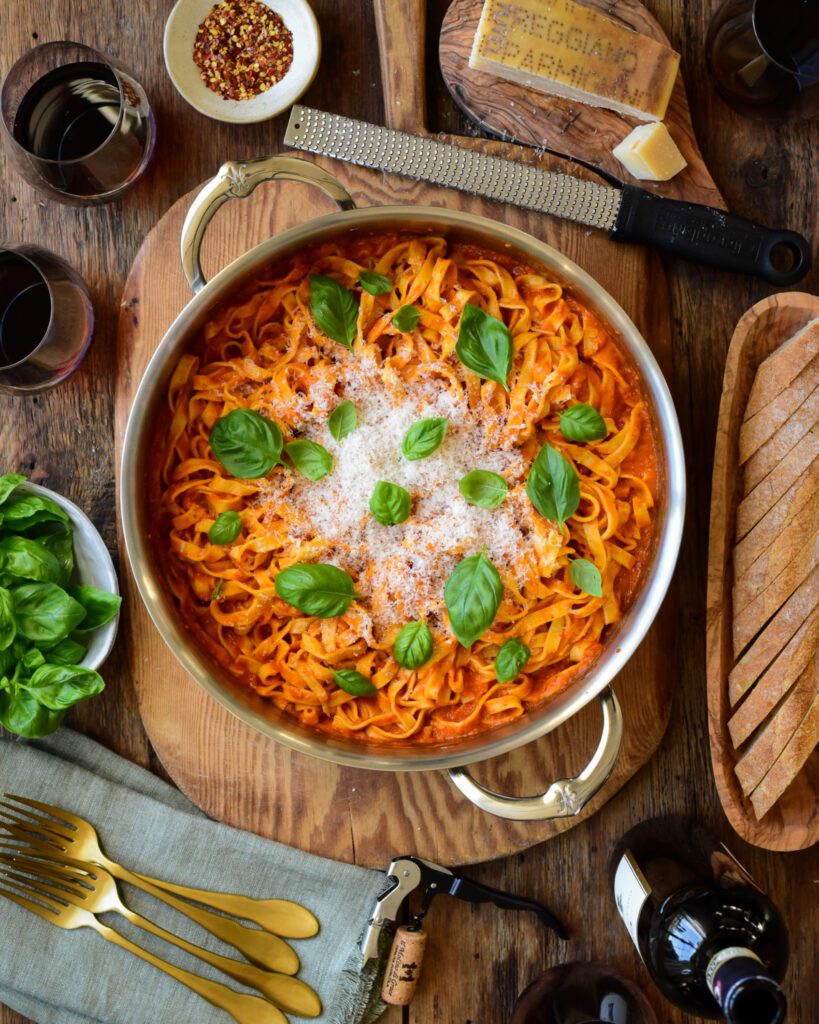 A two handled pot of homemade fresh pasta with a tomato sauce, basil and Parmesan. Surrounded with a a wooden bowl of sliced bread, a bowl full of fresh basil, a grater, fresh Parmesan, cutlery, napkins, wine glasses and wooden bowl of dried chillies.