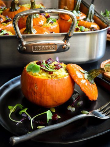 A baked mini pumpkin stuffed with rice with the lid leaning on it. A roaster in the background has more mini stuffed pumpkins.