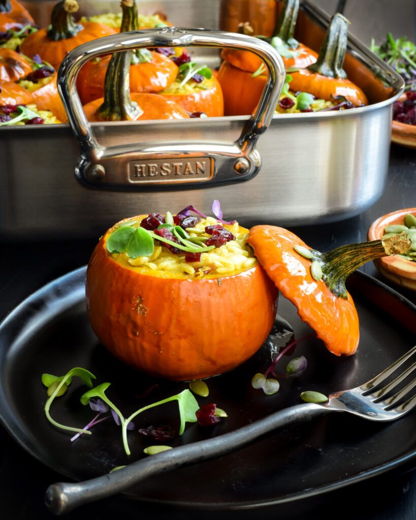 A baked mini pumpkin stuffed with rice with the lid leaning on it. A roaster in the background has more mini stuffed pumpkins.