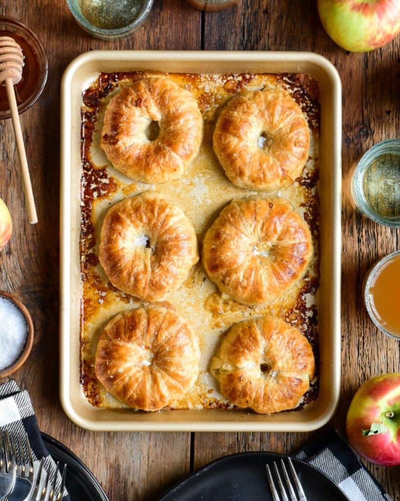 A close up of donut-like apple pies on a tray surrounded with a couple of glasses apples, honey and dishes.