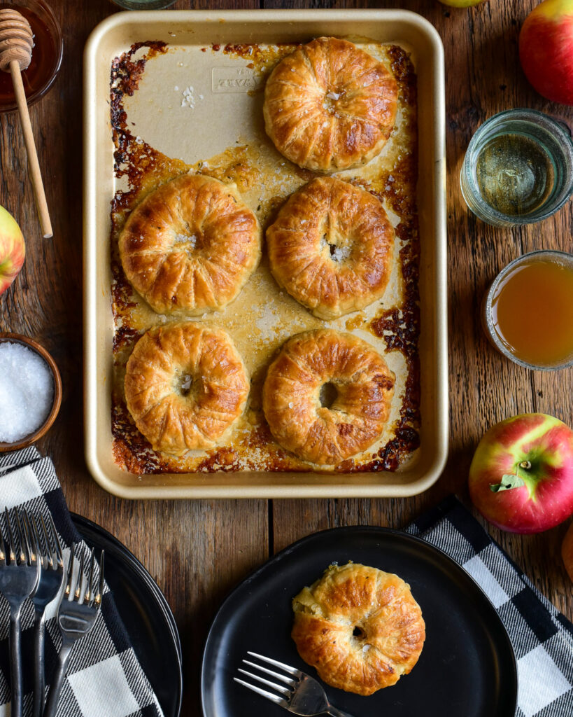 Five donut-like apple pies on a tray with one on a black plate. This tray is surrounded with a couple of glasses apples, honey and dishes.
