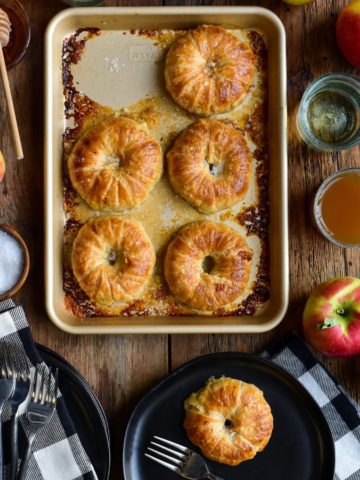 Five donut-like apple pies on a tray with one on a black plate. This tray is surrounded with a couple of glasses apples, honey and dishes.