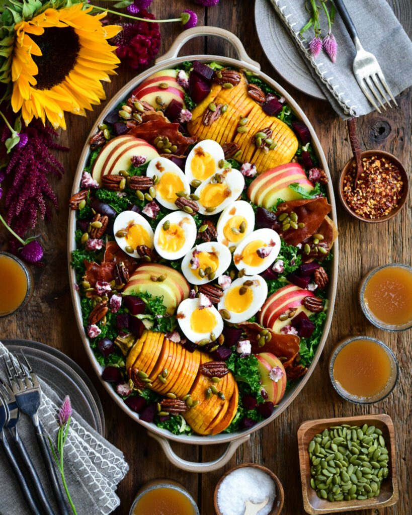An oval platter with a Fall Harvest Salad with Apple Cider Vinaigrette consisting of boiled eggs, hasselback squash, apple slices and kale. Surrounded with sunflowers, dishes, glasses with apple cider and other salad toppings.