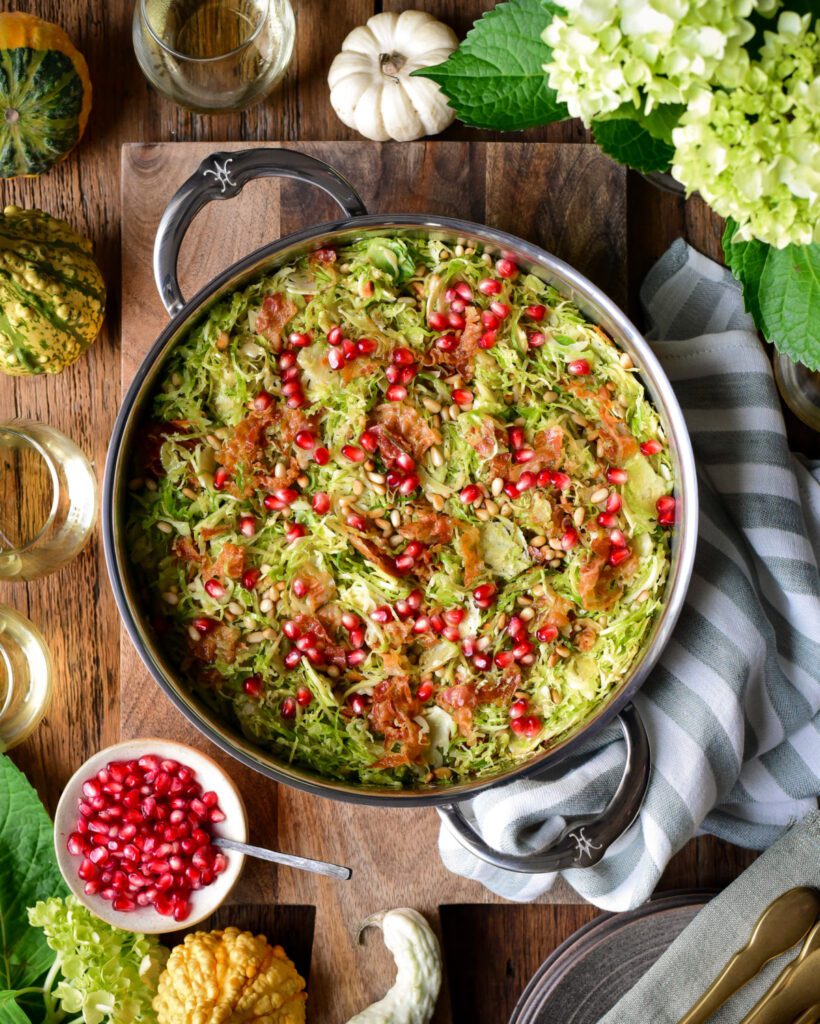 A top down image of brussels sprouts with pine nuts and pomegranates in a pan with two handles. Beside the pan is a tea towel and fall ornaments.