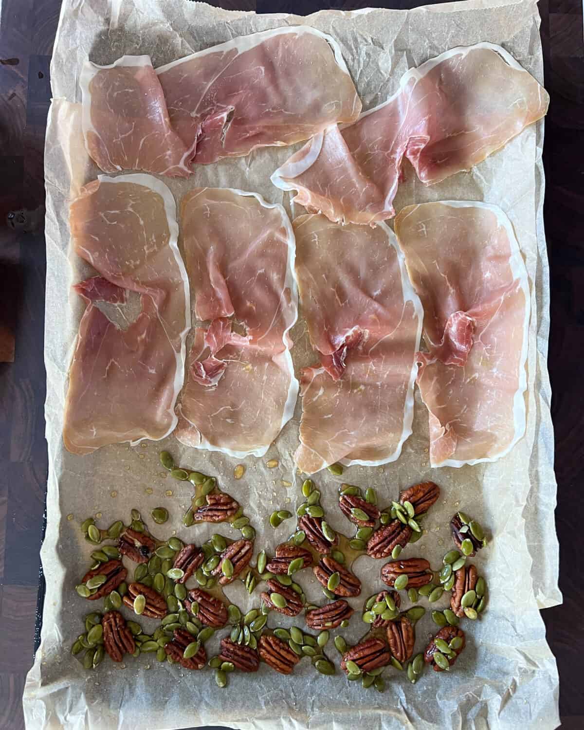 A sheet pan lined with parchment paper with prosciutto, nuts and seeds laid out to be roasted.