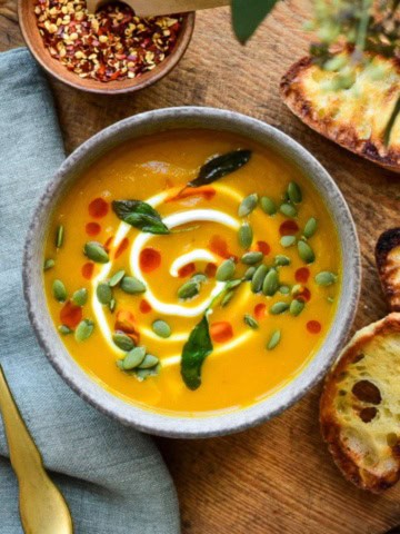 A bowl of Honeynut Squash Soup topped with a swirl of Greek yogurt, sprinkled with pepitas, chili oil and fried sage. Extra topping and three slices of baguette toast surround the bowl.