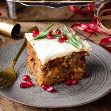 A corner slice of a Christmas carrot cake with cream cheese icing. Decorated with pomegranate arils and rosemary.